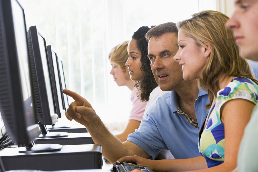 Four Students Sitting At Computer Terminals With Teacher Helping